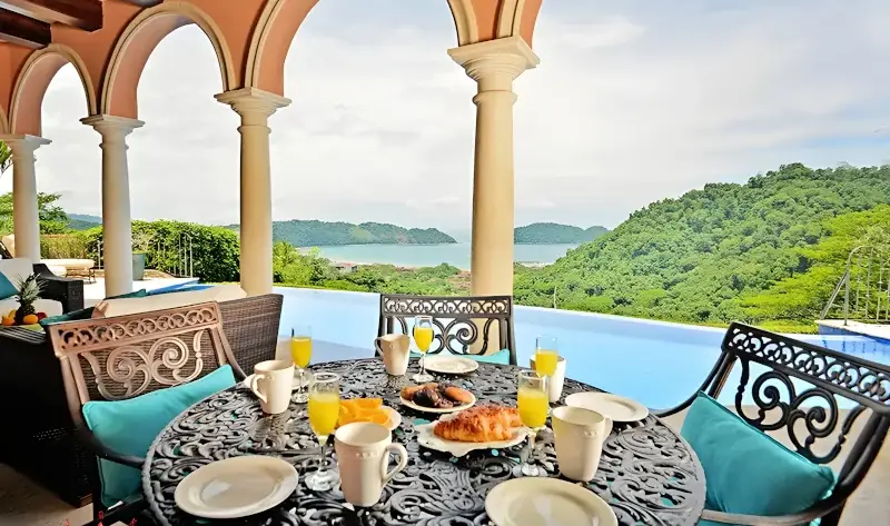 Outdoor dining area at Royale Hibiscus villa overlooking a private infinity pool with tropical mountain and ocean views, in Los Sueños Costa Rica